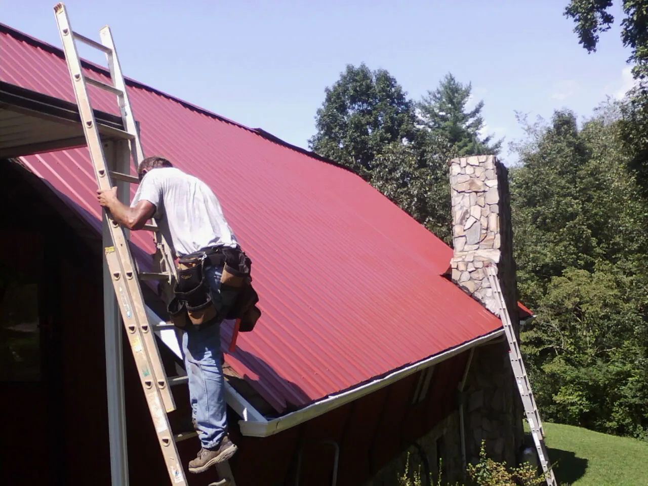 Roofer on a ladder beside a red metal roof and stone chimney — Soffit & Fascia Repair by Roofing Brothers in Dyersburg